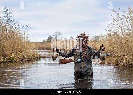 A hunter stands waist deep in water on a lake overgrown with reeds. He holds in his hands his prey - two shot ducks. The hunter is wearing a camouflag Stock Photo