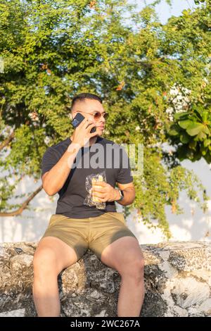 Casual man on phone call while holding a snack, seated outside with trees in the background. Stock Photo