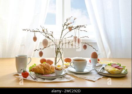 Breakfast with Easter decoration from natural colored eggs hanging on spring branches, coffee, croissant, bread roll and more on a wooden table, copy Stock Photo