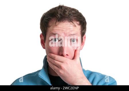 Frontal head portrait of a young brunette Caucasian man covering his mouth with his left hand while looking shocked at the camera. Stock Photo