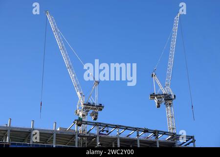 Two hoisting tower cranes on the top section being construction of modern high skyscraper  building against blue cloudless sky Stock Photo