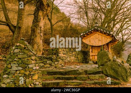Small wooden oriental building with tiled roof in front of rock wall at rural roadside park on winter evening Stock Photo