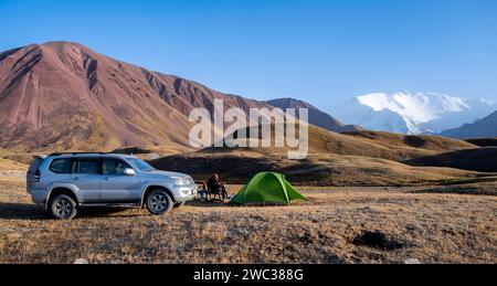 An off-road vehicle stands next to a pitched tent in a mountain landscape with snow-capped peaks in the background, Lenin Peak, Kyrgyzstan Stock Photo