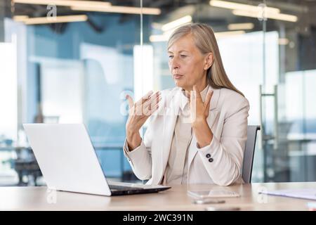 Focused senior businesswoman conversing on video call in modern office setting, expressing professionalism and engagement. Stock Photo