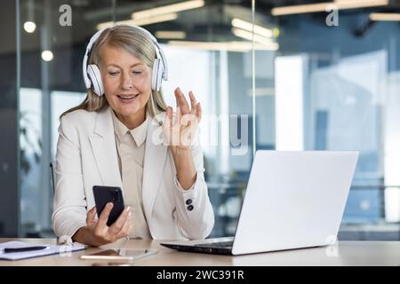 Mature businesswoman with headphones using smartphone and laptop, gesturing 'ok' in bright office setting, radiating positivity and professionalism. Stock Photo