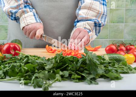 Selective focus on female hands cutting sweet pepper with a knife. Vegetables and greens in the foreground are blurred. Home cooking. Stock Photo