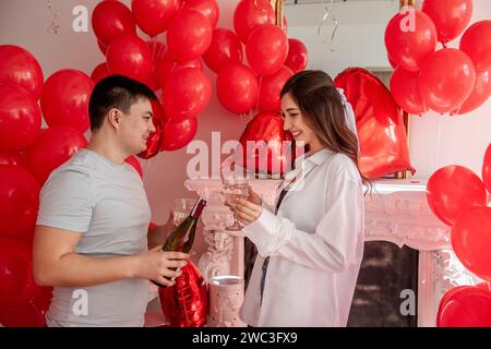 Joyous moment between young couple celebrating with toast Valentines day near red balloons, white fireplace. Woman laughing, holding glass, about to c Stock Photo