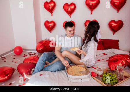 Joys of no-fuss Valentines Day celebration. Trend of combining fast food with romantic occasions. Cozy couple enjoying pizza from takeaway box, glasse Stock Photo
