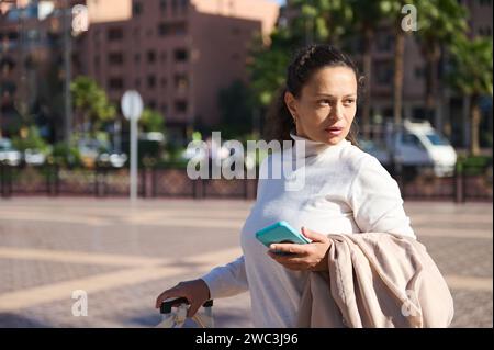 Thoughtful confident young adult multi ethnic woman holding mobile phone, looking aside, standing with her suitcase on the city street, waiting for ta Stock Photo