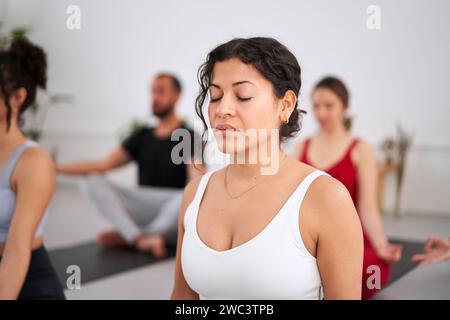 Close-up young Latina woman with eyes closed practising yoga exercises indoors. Group of people Stock Photo