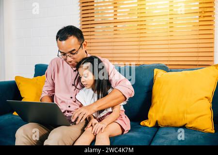 A smiling Asian father works from home on his laptop as his daughter learns on a computer for e Stock Photo