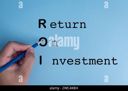Man hand holding a pencil to write ROI return on investment on blue background Stock Photo