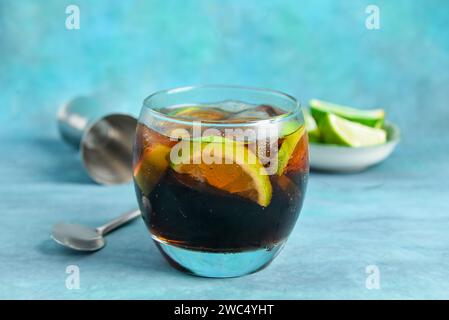 Glass of cold Cuba Libre cocktail and bowl with limes on blue background Stock Photo