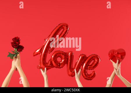 Female hands holding word LOVE made from balloons, gift box and roses on red background. Valentine's Day celebration Stock Photo