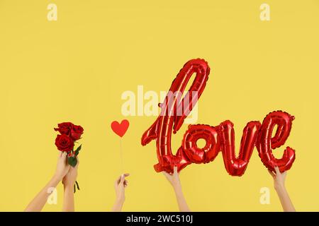 Female hands holding word LOVE made from balloons, paper heart and roses on yellow background. Valentine's Day celebration Stock Photo