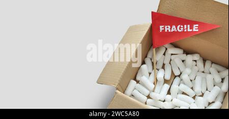 Opened cardboard packaging box with white polystyrene packing chips. Tiny paper flag with the warning 'Fragile' as a label. The concept of shipping fr Stock Photo