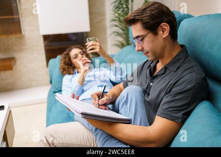 Focused young husband taking notes in notebook while sitting with wife on sofa Stock Photo