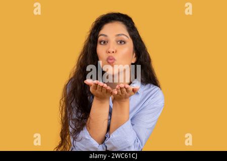 Charming young woman with long curly hair blowing a kiss, hands cupped in front Stock Photo