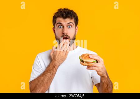 Portrait Of Funny Shocked Young Guy Eating Tasty Burger, Studio Stock Photo