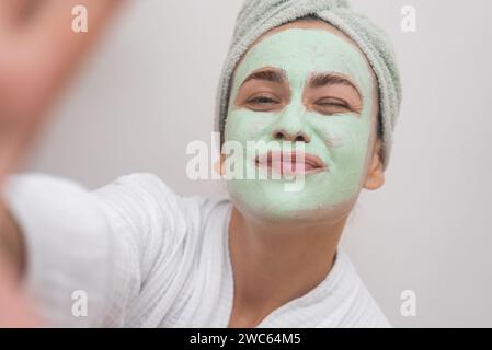 bathrobe-clad woman captures her joyful essence in a selfie with a clay mask, beaming at the camera. Stock Photo