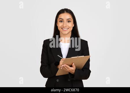 Happy female secretary, manager in black suit making notes at clipboard, standing isolated on gray studio background Stock Photo