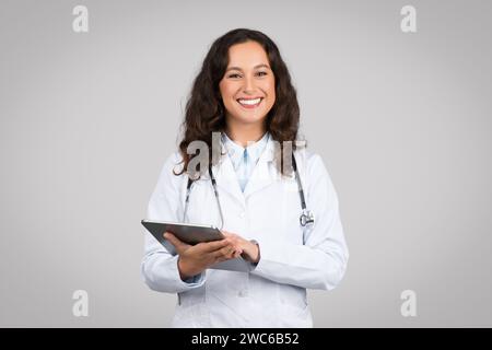 Positive woman in medical workwear doctor using digital tablet isolated on grey background, chatting with patient Stock Photo