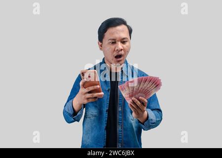 Young Asian man holding money and this phone showing shocked expression when looking to his money Stock Photo