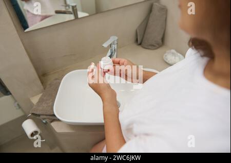 Details on the hands of a woman holding a white jar with moisturizer, standing in the home bathroom. View from above. The concept of healthy skin, bod Stock Photo