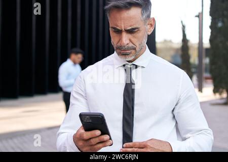 Attractive mature gray haired formal business man looks concentrated cell phone standing outdoor. Stock Photo