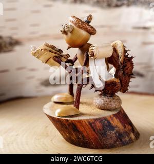 A little man made of acorns sits on a chestnut shell toilet and reads a book against a birch tree bark background. Square photo. Stock Photo