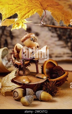 An acorn gnome reads a book while sitting on a stump under a sunny golden autumn leaf. There are many forest products around him. Thanksgiving theme. Stock Photo