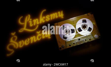 Card, with transparent audio cassette and yellow light text projection - Happy Holidays (text in Lithuanian) on dark background. Stock Photo