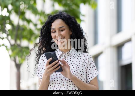Joyful woman with curly hair using smartphone outdoors. She exhibits happiness and connectivity in a casual urban setting. Stock Photo