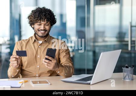A cheerful young man in a modern office setting using smartphone and credit card, possibly making an online transaction or shopping online. Stock Photo
