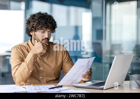 Thoughtful male professional reviews documents with deep focus. Office setting with laptop and papers captures a moment of analysis. Stock Photo