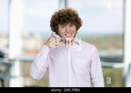 Handsome young man with curly hair shows call me gesture. Indoor window in the background. Stock Photo