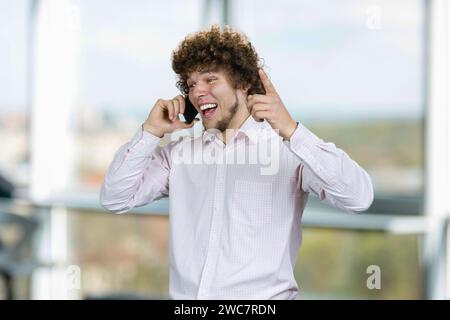 Portrait of a young happy man with curly hair talking on the phone rejoicing success. Indoor window in the background. Stock Photo