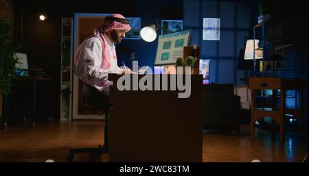 Professional muslim broker investor works with stock exchange valuation financial profit numbers. Arab stockholder in dimly lit apartment looking at market shares growth charts, taking notes Stock Photo