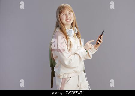 happy trendy teen girl in beige tracksuit with headphones using smartphone isolated on grey. Stock Photo