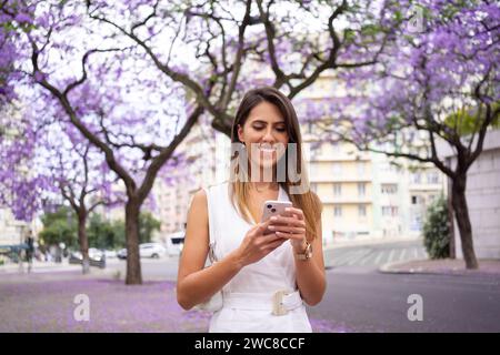 Smiling young woman in white dress with brown hair using smartphone outdoors. Confident businesswoman text messaging via cellphone near violet Jacaranda trees in city park Stock Photo