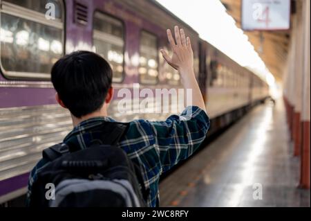 Back view image of a young Asian man waving his hand, say goodbye to someone on the train at a railway station. People and transportation concepts Stock Photo