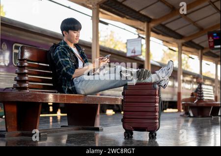 A happy, handsome Asian man is using his phone, chatting with his friends or scrolling on social media while sitting on a bench, waiting for his train Stock Photo