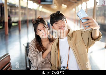 Cute and happy young Asian couple taking selfies with a smartphone while waiting for the train at a railway station together, excited for their summer Stock Photo