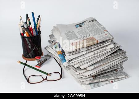 stacks of newspapers that need to be read to get information on business and financial news, plus stationery and glasses on the table Stock Photo