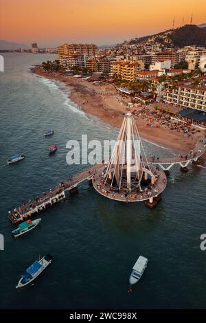 Closer view of Los Muertos Beach Pier at sunset with the sky illuminating at golden hour. Boats below and people walking on path. Stock Photo