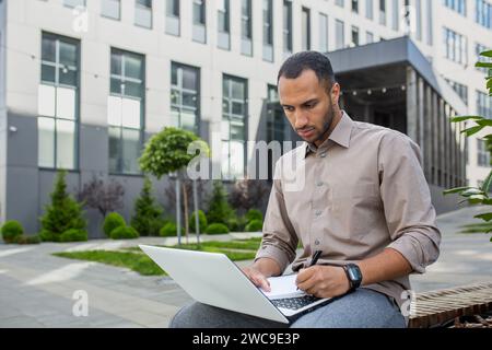 Focused young man using a laptop outdoors with a modern building backdrop, embodying mobile work and concentration. Stock Photo