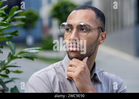 Thoughtful young man in glasses looking away, hand on chin, embodying contemplation and decision-making in an urban setting. Stock Photo
