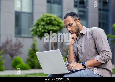 Focused man with glasses working on laptop outdoors, showing concentration and determination in a casual urban setting. Stock Photo