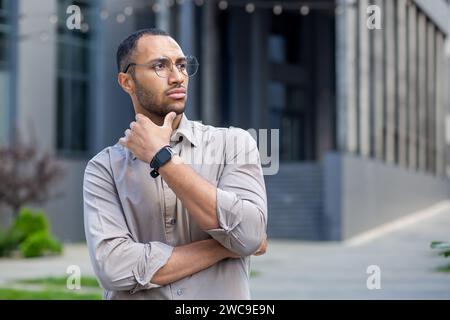 Pensive young man with glasses and smartwatch in urban setting, hand on chin, contemplating, with a focused expression. Stock Photo