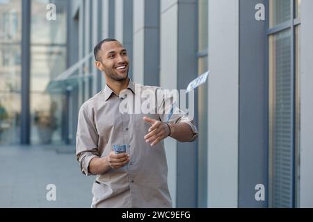Smiling man casually dressed in beige shirt outside, extending a handshake, holding phone and money, conveying trust and friendliness. Stock Photo